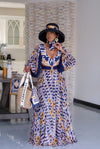 Model wearing a vibrant, patterned maxi dress featuring a bold mix of geometric and floral prints from the Tshwane Resort Collection. The look is elevated with a deep V-neckline, statement sleeves, and a waistbelt with a gold FRS buckle that adds definition. Styled with a wide-brimmed Fabrosanz hat and accessorized with a Fabrosanz bag for a polished resort-ready aesthetic.