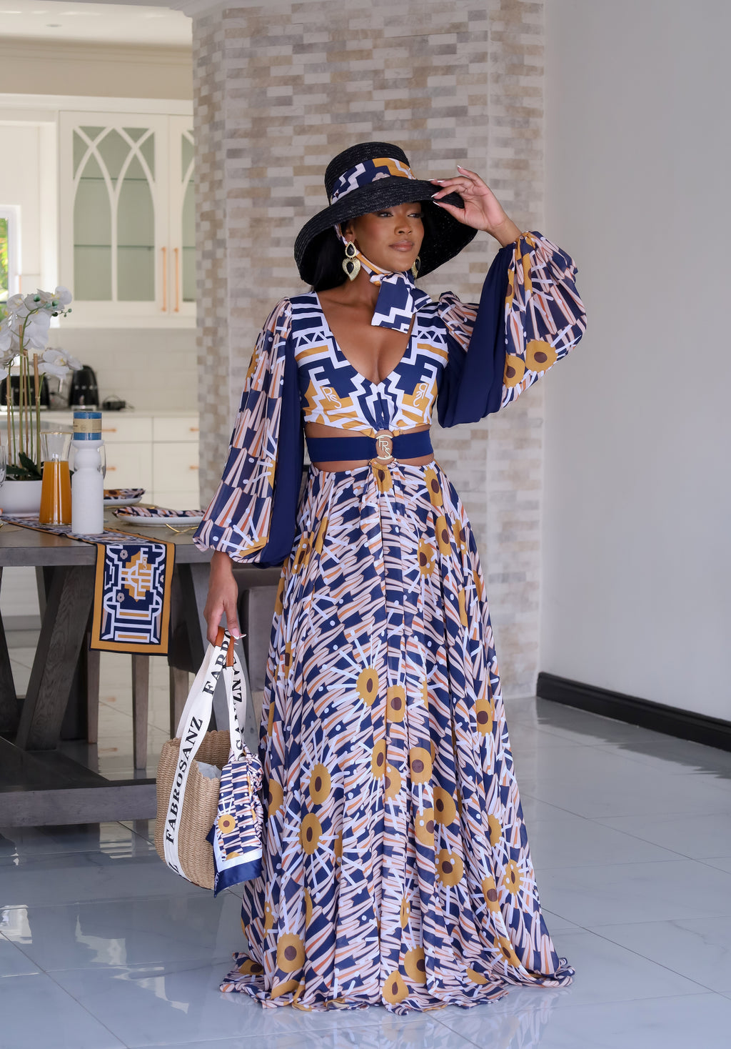 Model wearing a vibrant, patterned maxi dress featuring a bold mix of geometric and floral prints from the Tshwane Resort Collection. The look is elevated with a deep V-neckline, statement sleeves, and a waistbelt with a gold FRS buckle that adds definition. Styled with a wide-brimmed Fabrosanz hat and accessorized with a Fabrosanz bag for a polished resort-ready aesthetic.