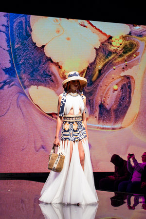 A model on the runway wearing a stunning geometric-patterned dress with a dramatic white tulle skirt and a bold navy, orange, and white tribal-inspired bodice. The look is completed with a wide-brim straw hat and a woven handbag, set against a vivid artistic background.